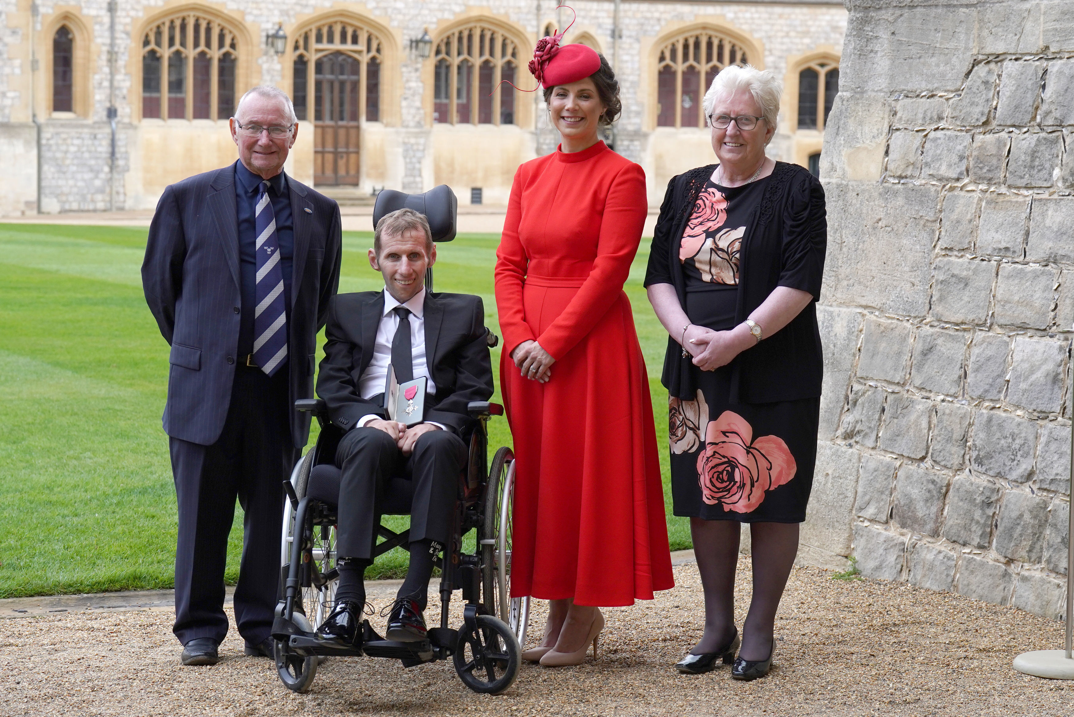 A group photo of four people at an investiture ceremony at Windsor Castle; one recipient is in a wheelchair.